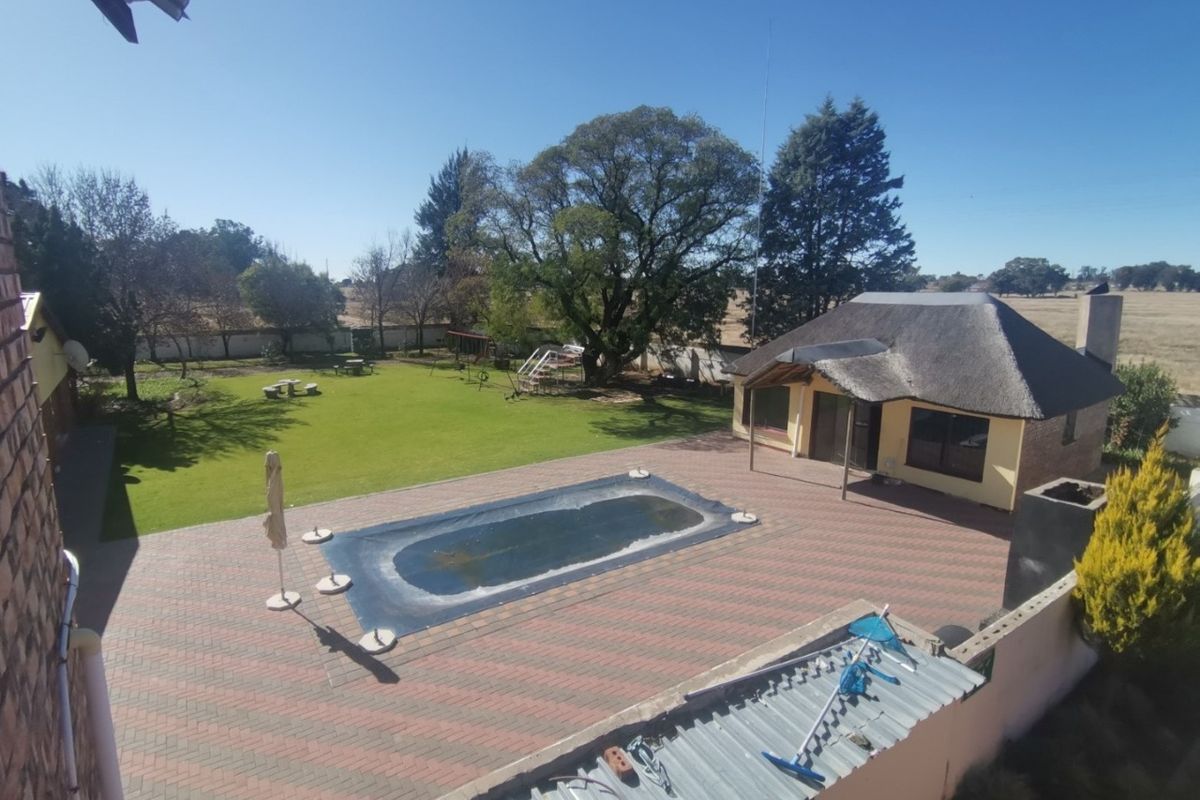 Aerial view of a small holding featuring a swimming pool, paved patio area, and a separate thatched-roof building set within a spacious garden.