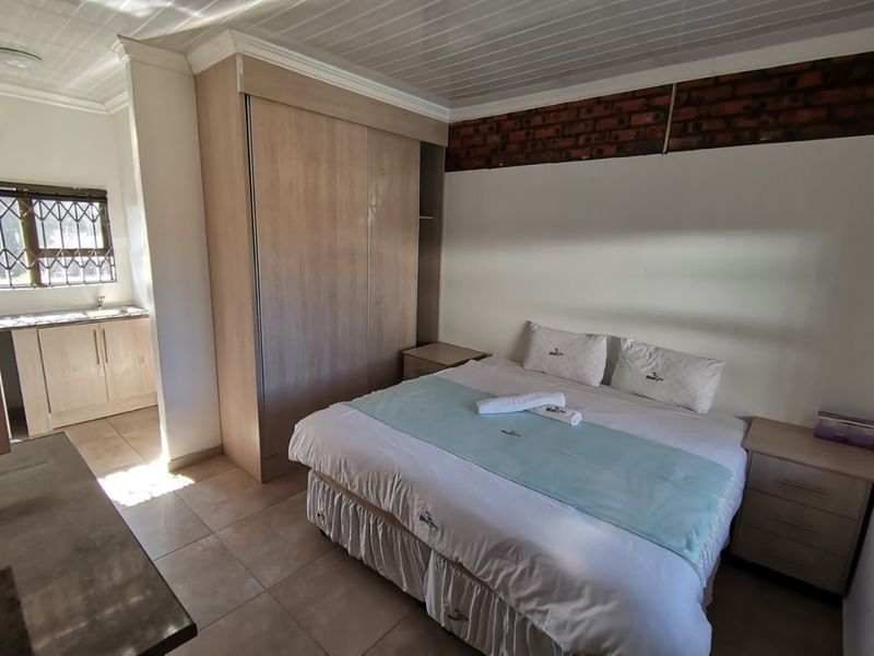 A bedroom featuring built-in cupboards, tiled flooring, and an exposed brick accent wall, with an adjoining small kitchenette area visible.
