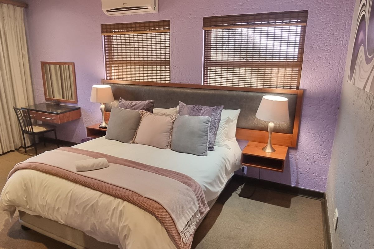 A bedroom featuring a built-in headboard with integrated bedside tables, wall-mounted air conditioning unit, and dual windows with bamboo blinds.