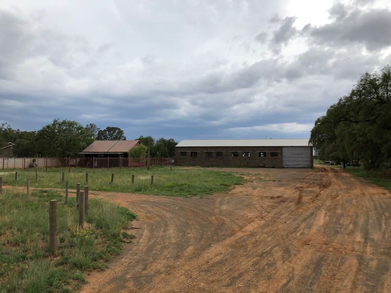 Exterior view of a small holding property featuring a large brick outbuilding, metal roofing, and an unpaved driveway.