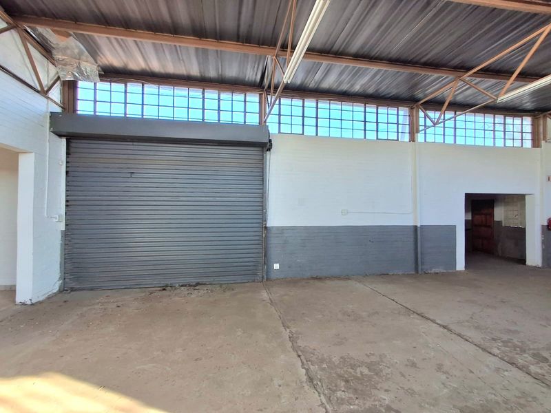 Spacious commercial utility area featuring a large roller shutter door, high ceilings with exposed steel trusses, and ample natural light from clerestory windows.