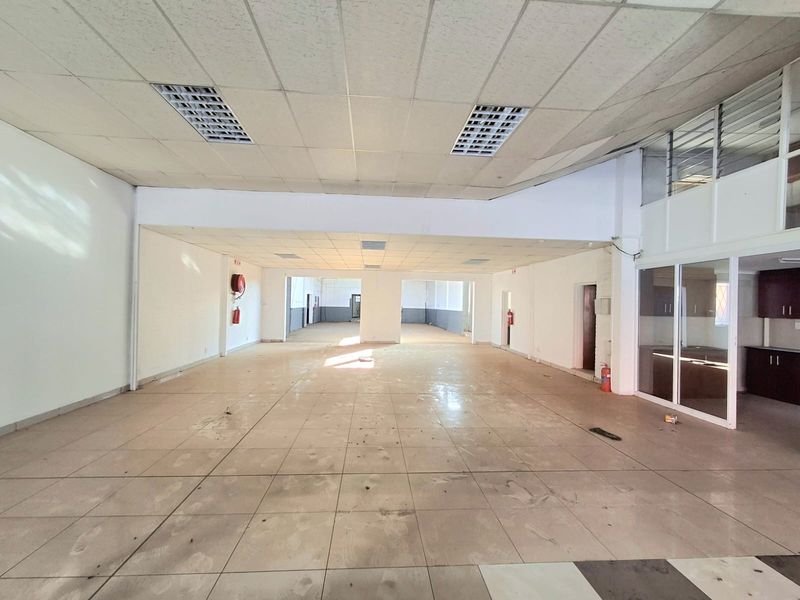 Spacious open-plan commercial area featuring tiled flooring, suspended ceilings with recessed lighting, and a glass-partitioned office or storage section.