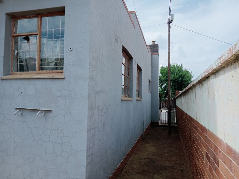 Side exterior view of the commercial building showing a narrow walkway, brick boundary wall, and weathered plastered walls with steel-framed windows.