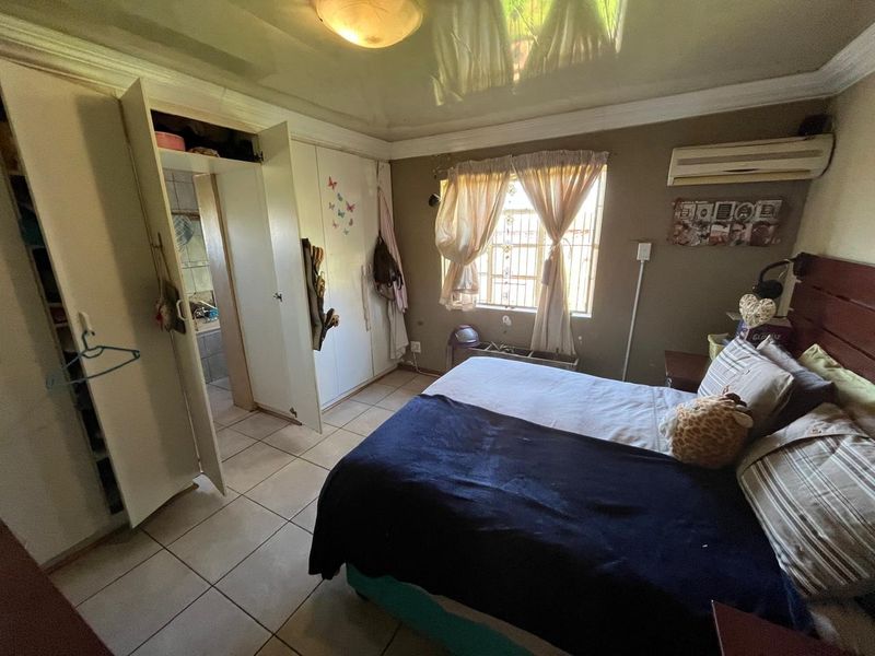 A bedroom featuring tiled flooring, built-in cupboards, and an en-suite bathroom. The room includes an air conditioning unit and security bars on the window.