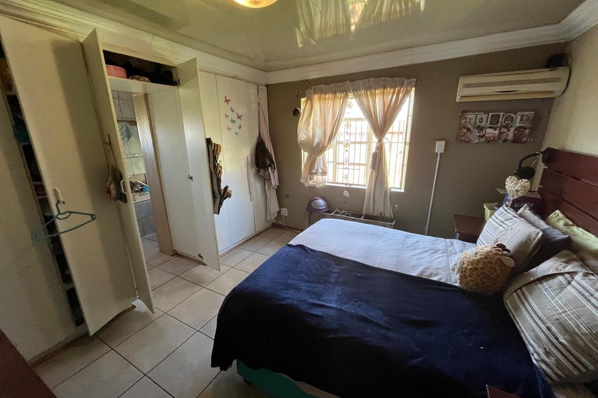 A bedroom featuring tiled flooring, built-in cupboards, and an en-suite bathroom. The room includes an air conditioning unit and security bars on the window.