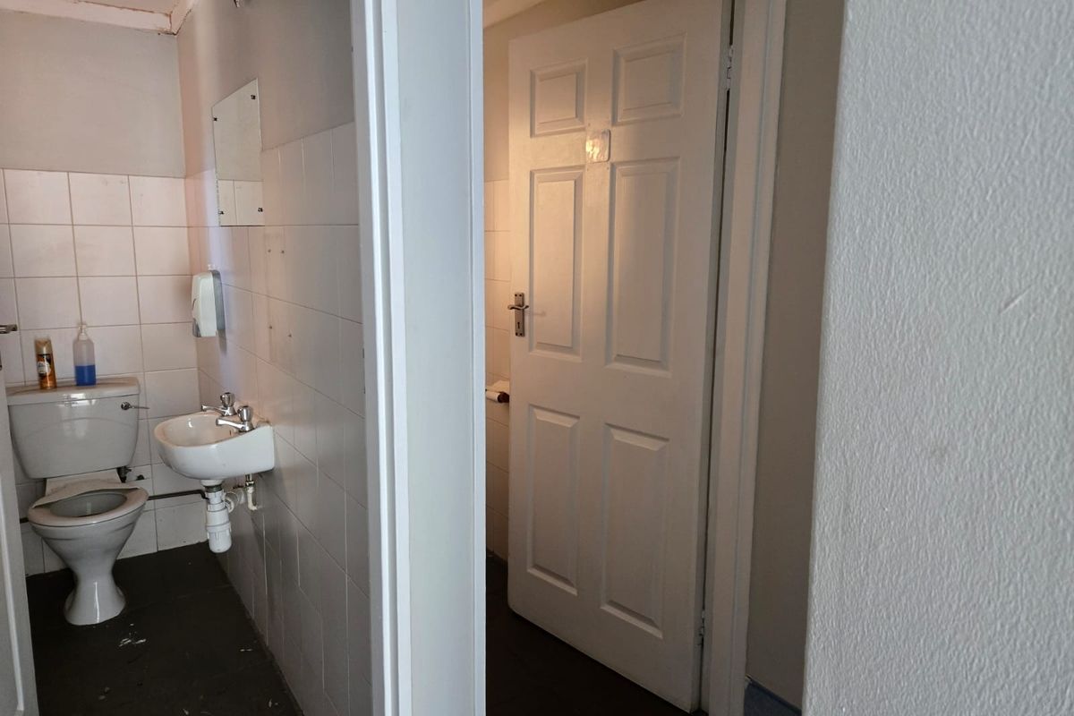 A functional commercial bathroom featuring a wall-mounted basin, toilet, and tiled walls, requiring some maintenance and cosmetic updates.