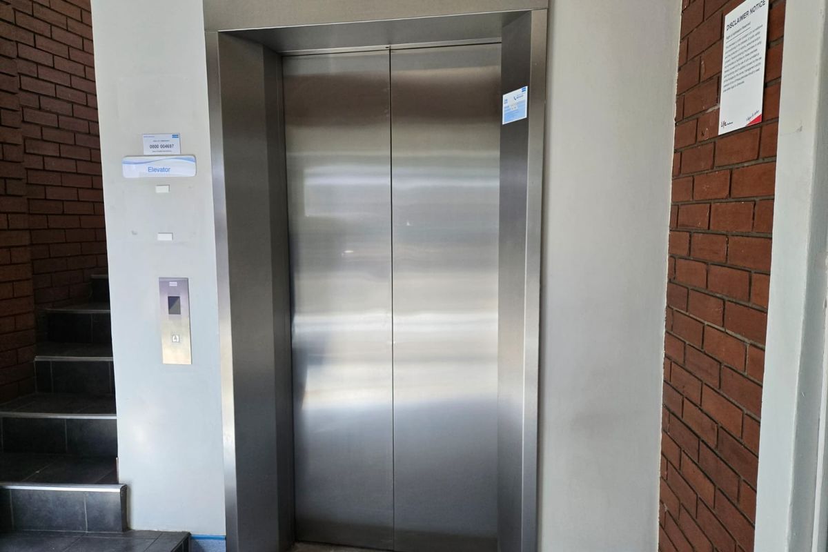A commercial building hallway featuring a stainless steel elevator entrance, adjacent brick wall detailing, and a tiled staircase.