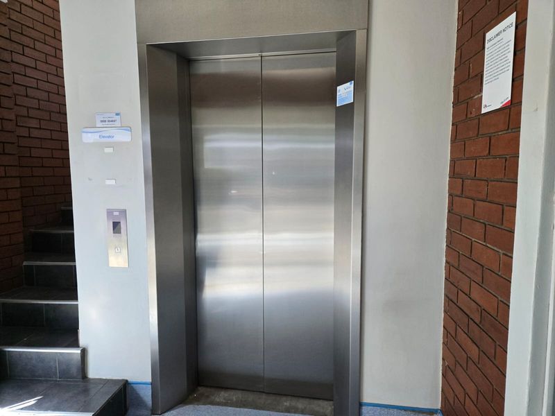 A commercial building hallway featuring a stainless steel elevator entrance, adjacent brick wall detailing, and a tiled staircase.