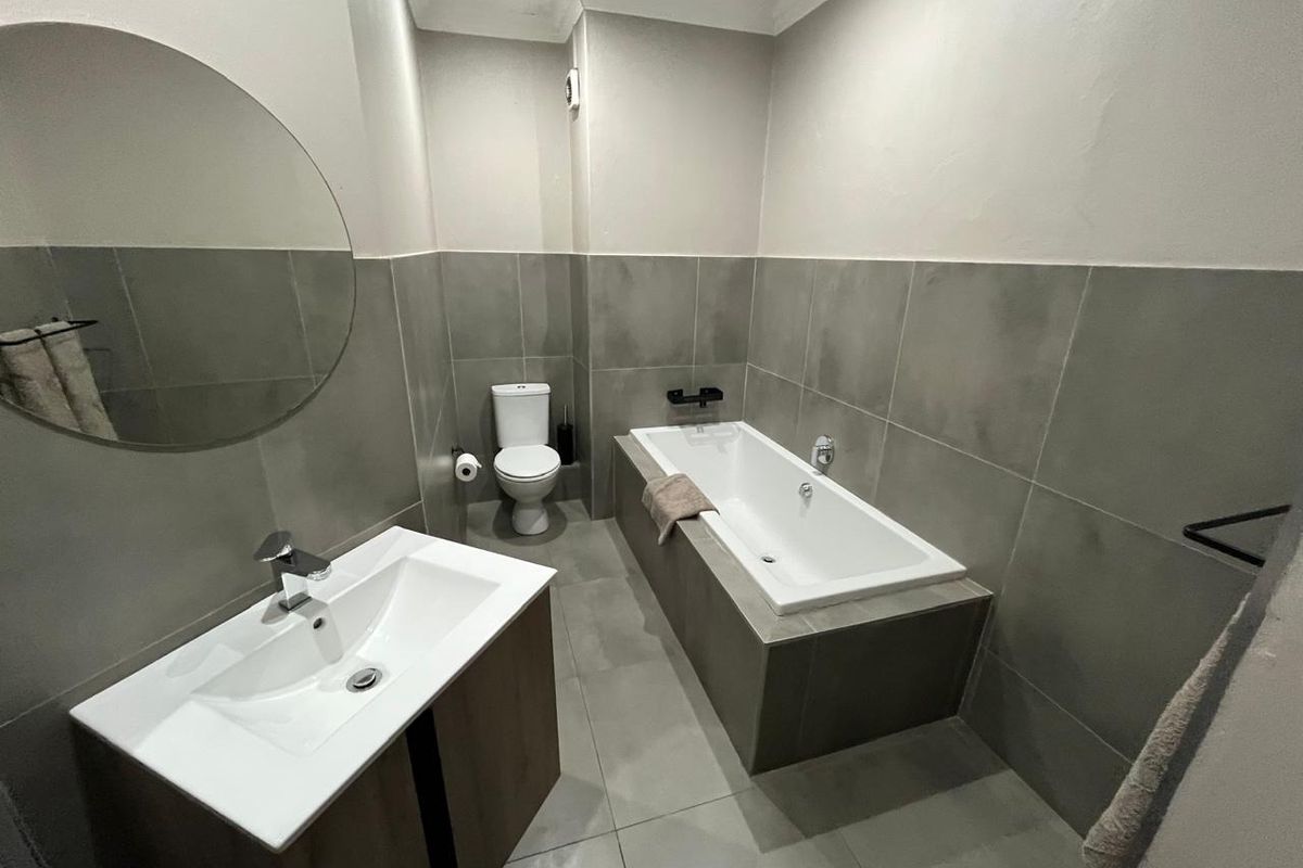 Modern bathroom featuring a built-in bathtub, vanity with basin, and toilet, finished with neutral grey wall and floor tiles.