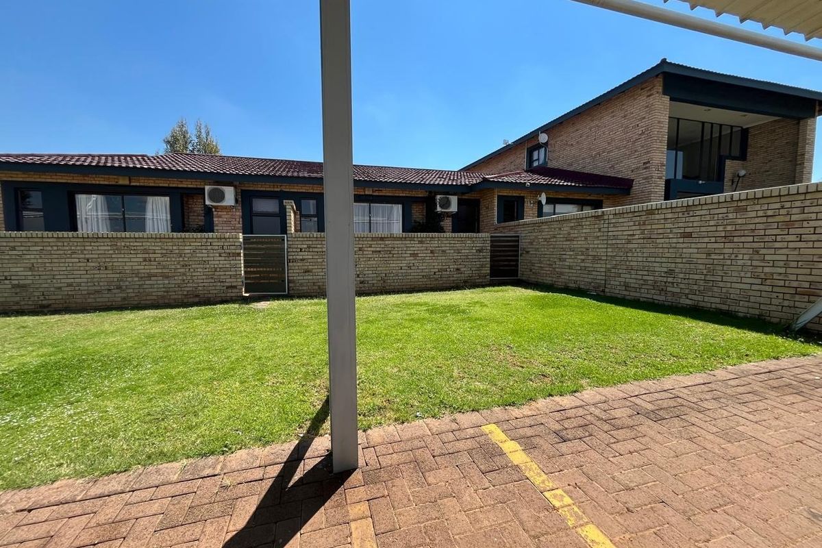 A rear exterior view of the guest house featuring a paved parking area, a well-maintained lawn, and a brick boundary wall.
