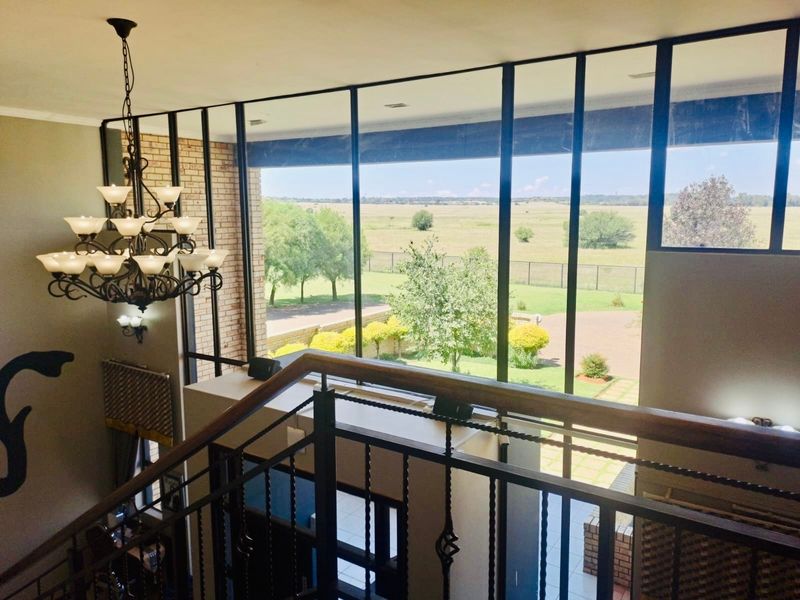 An elegant double-volume entrance hall featuring a wrought iron staircase, grand chandelier, and expansive floor-to-ceiling windows offering scenic views of the surrounding landscape.