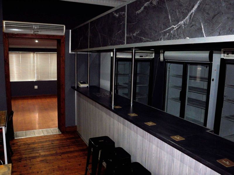 A commercial bar area featuring a dark-toned counter, overhead storage, integrated glass-door refrigeration units, and polished timber flooring.