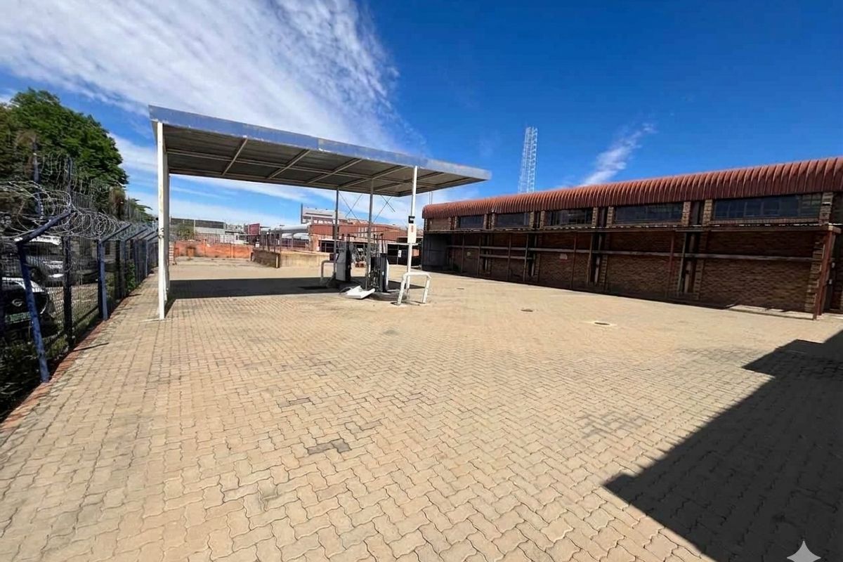 Commercial property exterior featuring a paved forecourt, fuel pump canopy, and adjacent brick building with security fencing.
