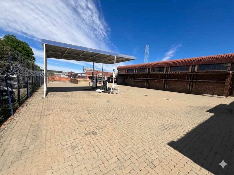 Commercial property exterior featuring a paved forecourt, fuel pump canopy, and adjacent brick building with security fencing.