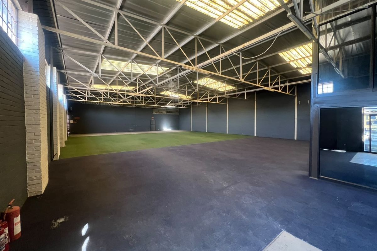 Spacious open-plan commercial warehouse area featuring exposed steel roof trusses, natural light from skylights, painted brick walls, and a combination of rubberized and turf flooring.