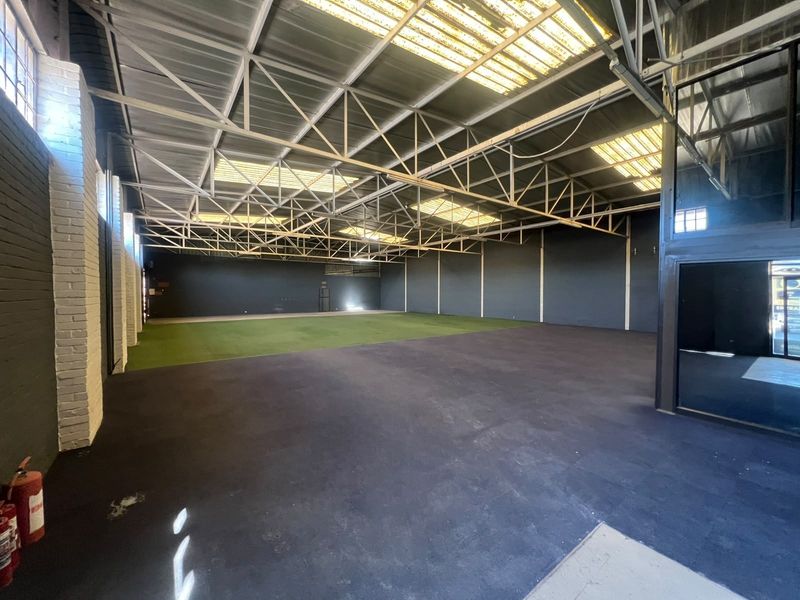 Spacious open-plan commercial warehouse area featuring exposed steel roof trusses, natural light from skylights, painted brick walls, and a combination of rubberized and turf flooring.