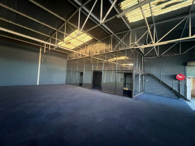Spacious commercial warehouse area featuring exposed steel roof trusses, a mezzanine level with glass partitioning, and a metal staircase.