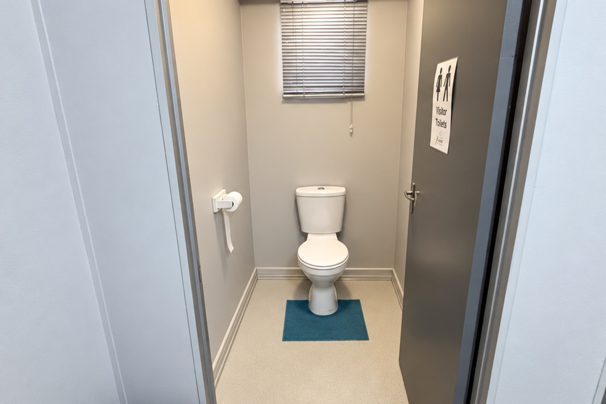 A clean and functional visitor restroom featuring a standard toilet, neutral wall finishes, and a window with a venetian blind.