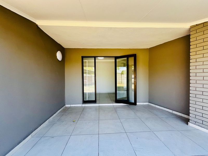 A clean, covered patio area featuring large format floor tiles, a brick feature wall, and double glass doors leading into the home.