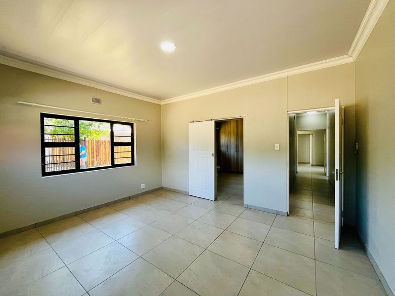 A spacious, empty bedroom featuring neutral tiled flooring, recessed lighting, coving, and built-in cupboards with convenient access to an adjoining room.