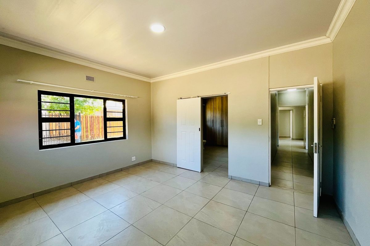 A spacious, empty bedroom featuring neutral tiled flooring, recessed lighting, coving, and built-in cupboards with convenient access to an adjoining room.