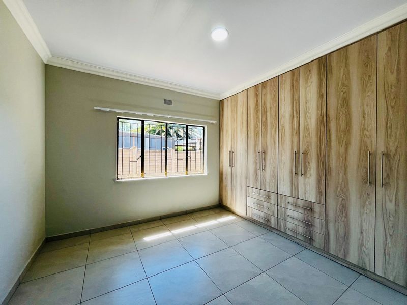 A bright, empty bedroom featuring modern wood-finish built-in cupboards, neutral tiled flooring, and security bars on the window.