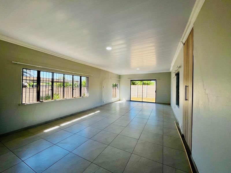 A spacious, empty living area featuring neutral tiled flooring, recessed lighting, and large windows with security bars, leading out to a rear outdoor space.
