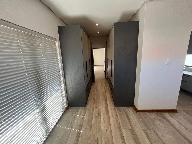 A modern hallway featuring extensive built-in dark cabinetry, light wood-look laminate flooring, and large windows with fitted venetian blinds.