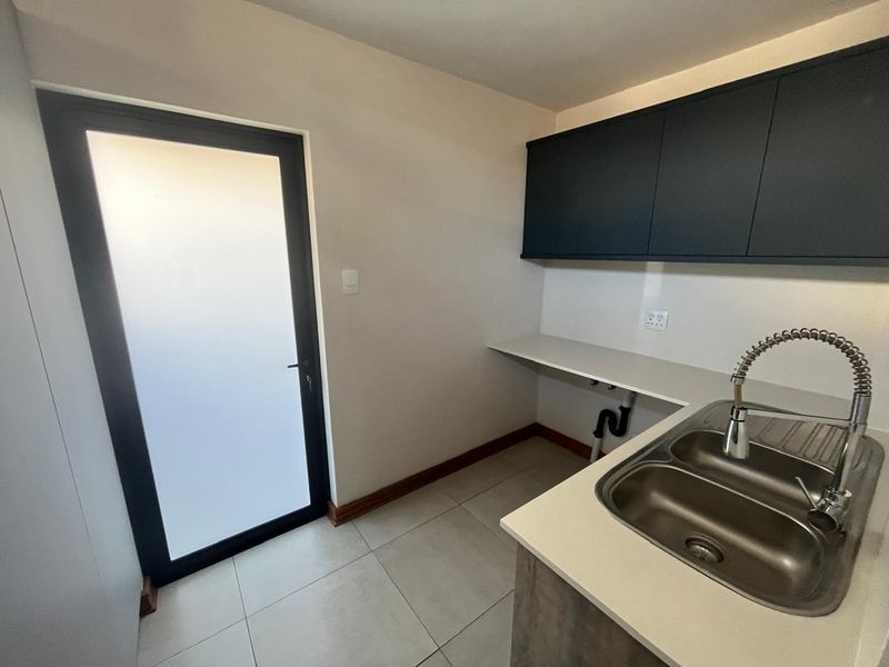 A clean, modern scullery featuring a double bowl stainless steel sink, gooseneck tap, ample counter space, dark overhead cabinetry, and a frosted glass exterior door.