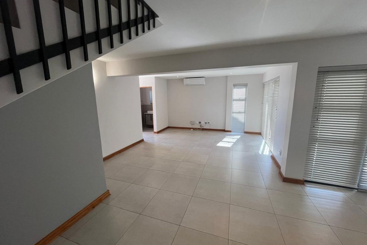 Spacious open-plan living area featuring neutral tiled flooring, a staircase, air conditioning, and large windows with fitted blinds.