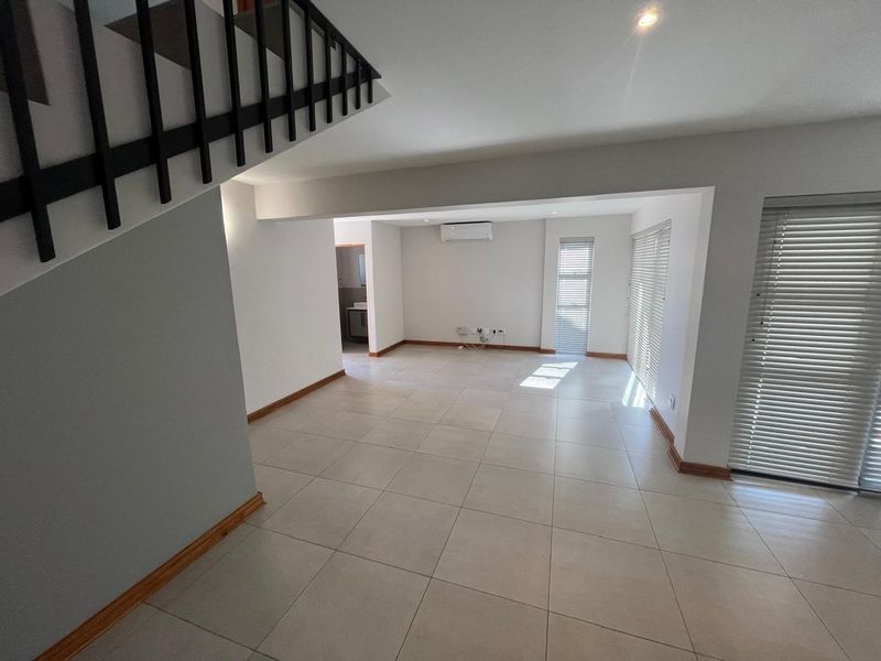 Spacious open-plan living area featuring neutral tiled flooring, a staircase, air conditioning, and large windows with fitted blinds.