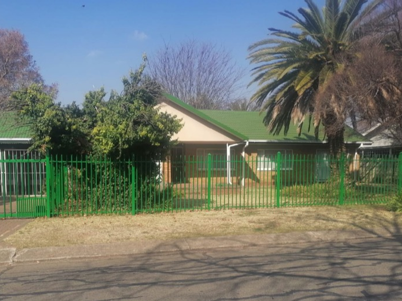 Front view of a single-story brick house featuring a green tiled roof and secure perimeter fencing.