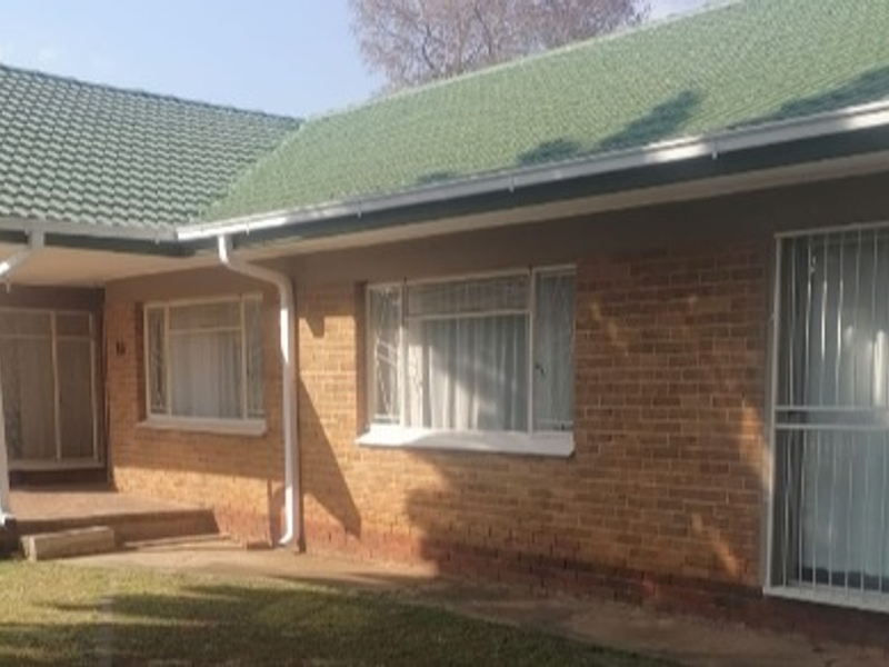 Front elevation of a single-story brick house featuring a green tiled roof, security bars on windows, and a paved entrance area.