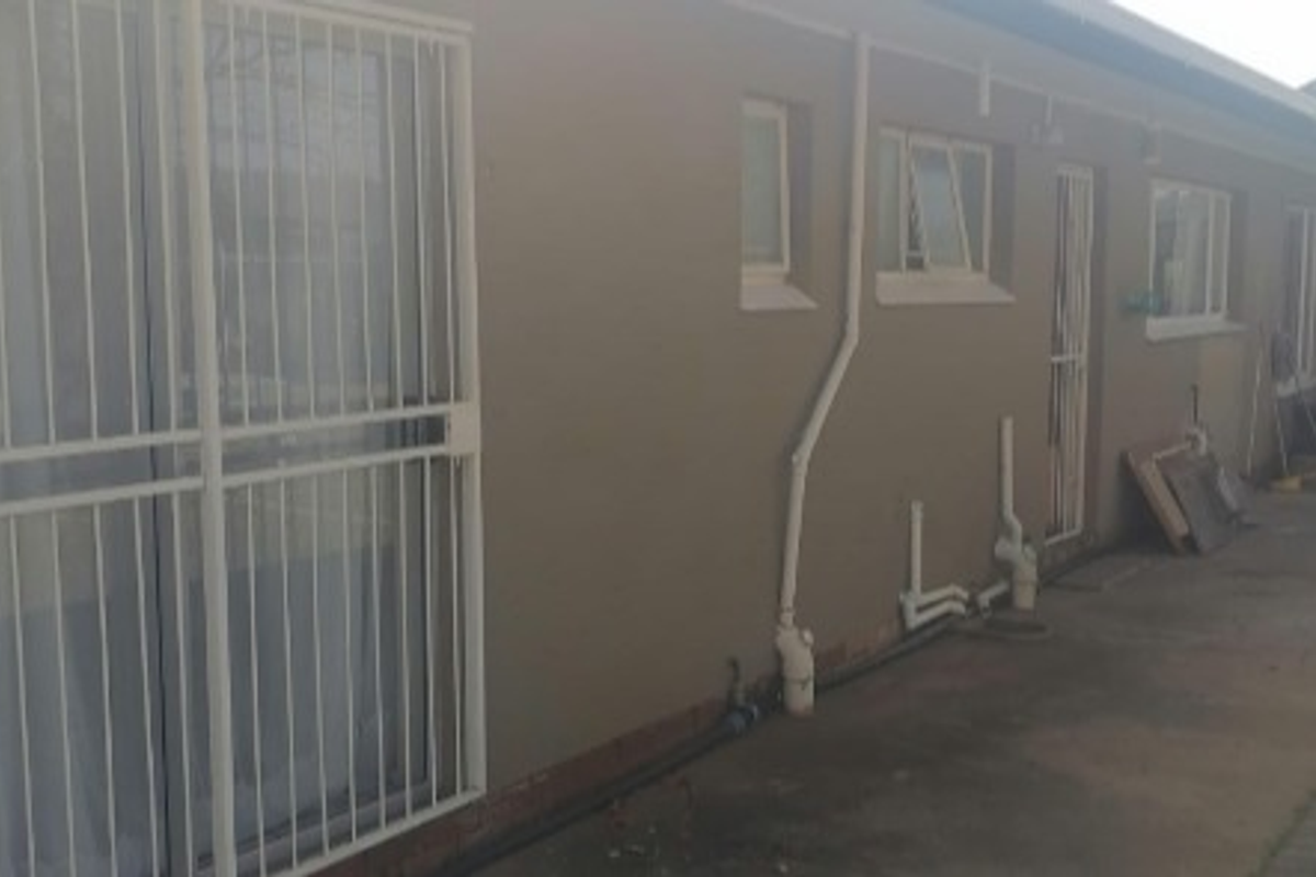 Rear exterior view of the house featuring security gates on sliding doors, casement windows, and exposed exterior plumbing along the concrete pathway.