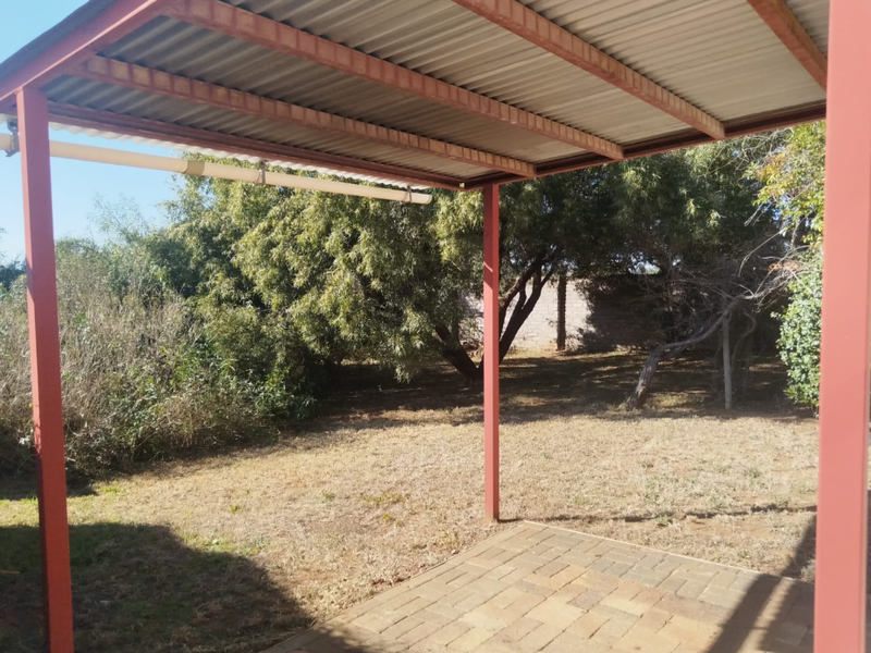 A covered outdoor patio area featuring a steel-framed structure with a corrugated metal roof, overlooking a spacious garden.