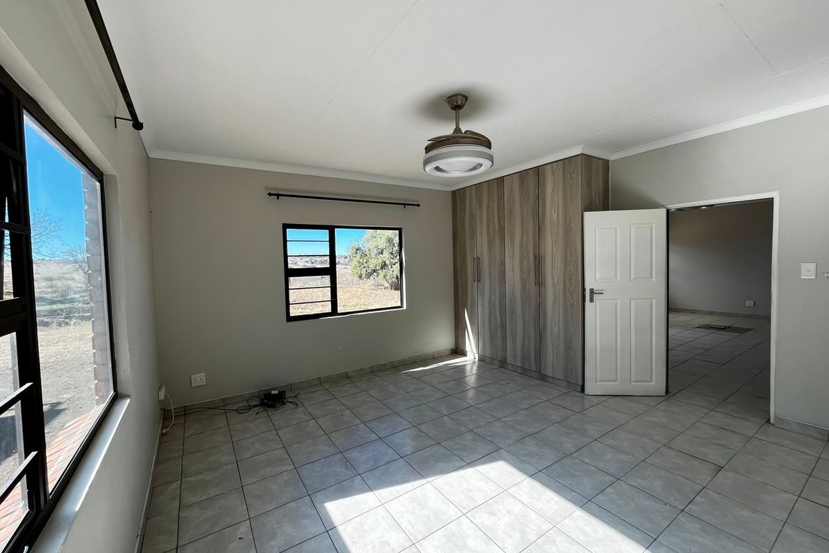 A bright, empty bedroom featuring neutral tiled flooring, built-in wooden cupboards, a ceiling fan, and large windows with security bars.