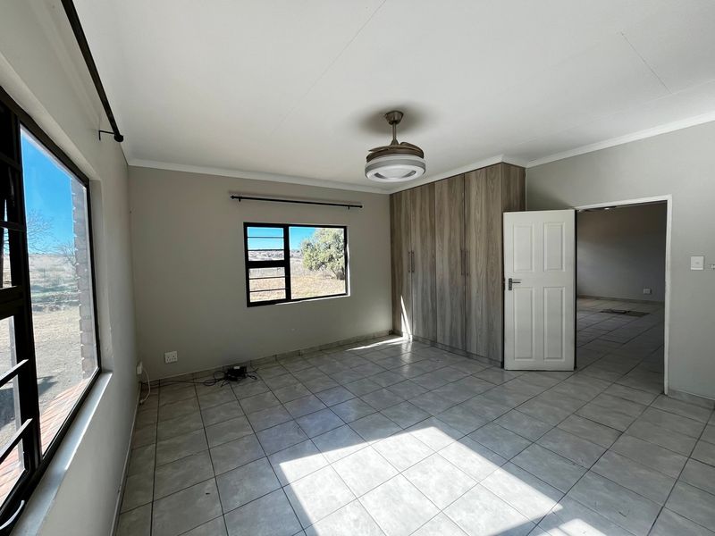 A bright, empty bedroom featuring neutral tiled flooring, built-in wooden cupboards, a ceiling fan, and large windows with security bars.