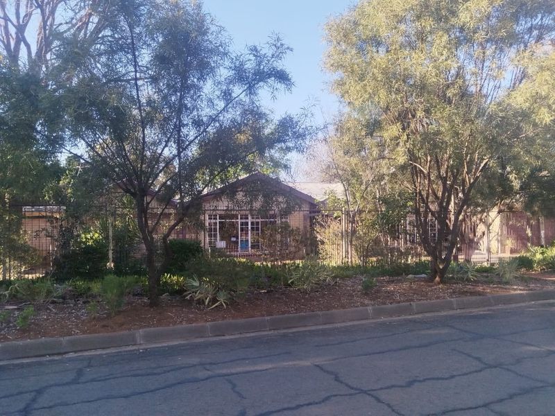 Front view of a single-story brick house featuring a tiled roof and secure metal perimeter fencing, set behind established trees and garden beds.