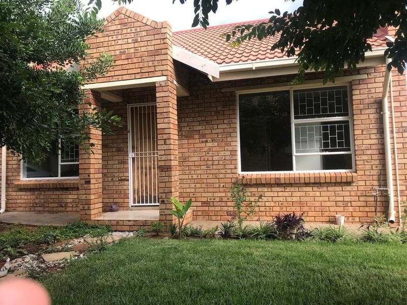 Front elevation of a face brick townhouse featuring a tiled roof, security gate, and burglar bars with a small front garden and paved walkway.