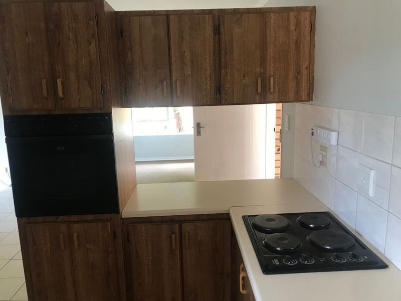 A compact kitchen featuring dated wood-look cabinetry, a built-in oven, electric hob, and tiled splashback, offering potential for a modern renovation.