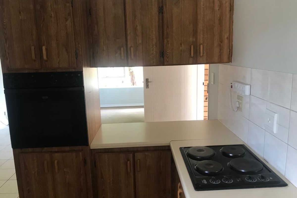 A compact kitchen featuring dated wood-look cabinetry, a built-in oven, electric hob, and tiled splashback, offering potential for a modern renovation.