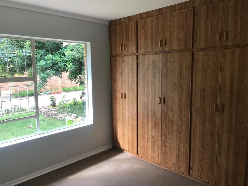 A bright, empty bedroom featuring extensive built-in wooden cupboards and a large window overlooking the garden.