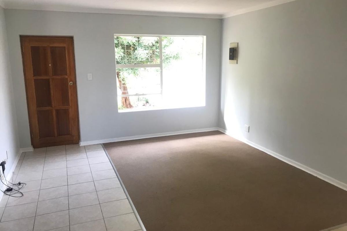 A bright, empty living room featuring a combination of tiled and carpeted flooring, a wooden entrance door, and a large window with security bars.