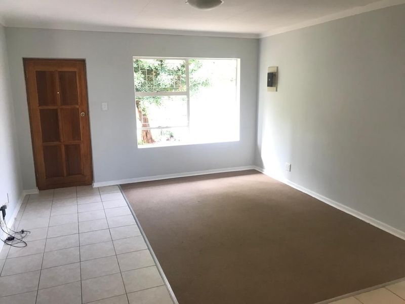 A bright, empty living room featuring a combination of tiled and carpeted flooring, a wooden entrance door, and a large window with security bars.