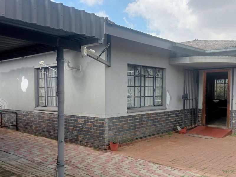Front exterior view of a single-story house featuring a brick and rendered facade, metal roof, security bars, and a paved driveway with a carport.