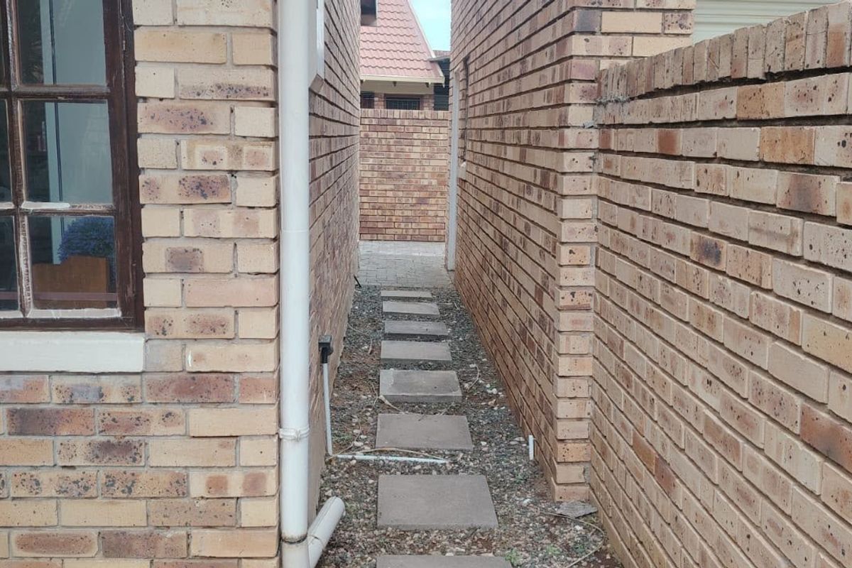 A narrow side passage between two face brick townhouse walls featuring a paved walkway and a boundary wall.
