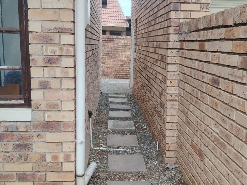 A narrow side passage between two face brick townhouse walls featuring a paved walkway and a boundary wall.