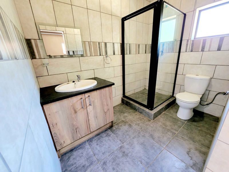 A clean, modern bathroom featuring a vanity cabinet with basin, a glass-enclosed shower, and a toilet, finished with neutral wall and floor tiling.