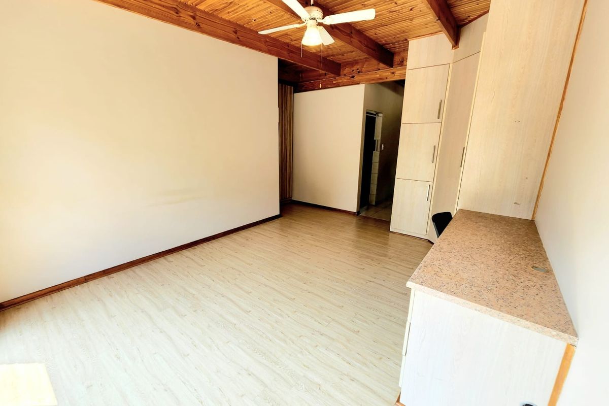 An empty bedroom featuring a rustic exposed timber beam ceiling, ceiling fan, laminate flooring, and built-in storage cupboards with an integrated desk area.
