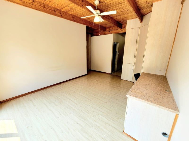 An empty bedroom featuring a rustic exposed timber beam ceiling, ceiling fan, laminate flooring, and built-in storage cupboards with an integrated desk area.